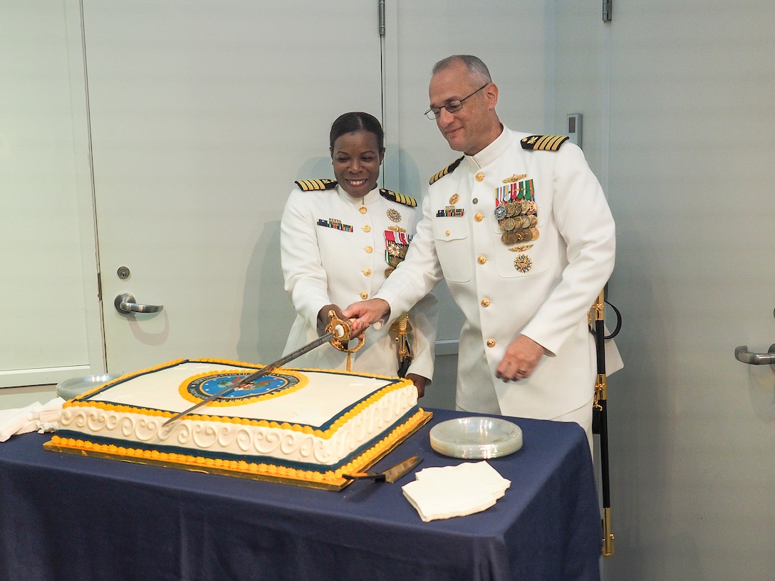 Two Navy officers cut a cake with a sword.
