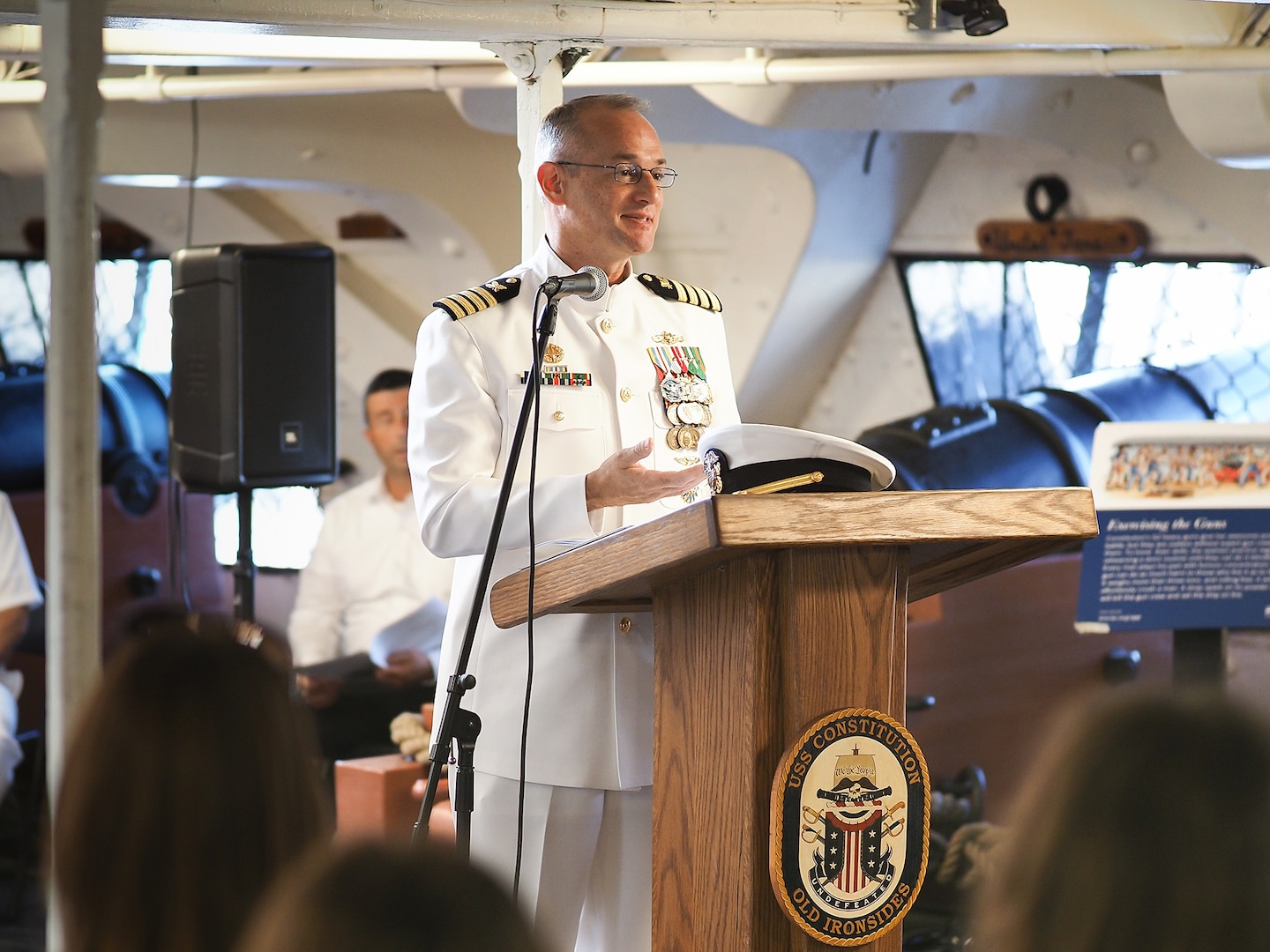 A Navy officer in a white uniform speaks from a podium.