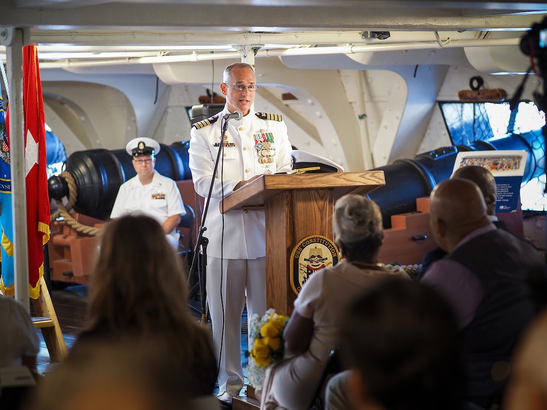 A Navy officer in white uniform speaks from a podium