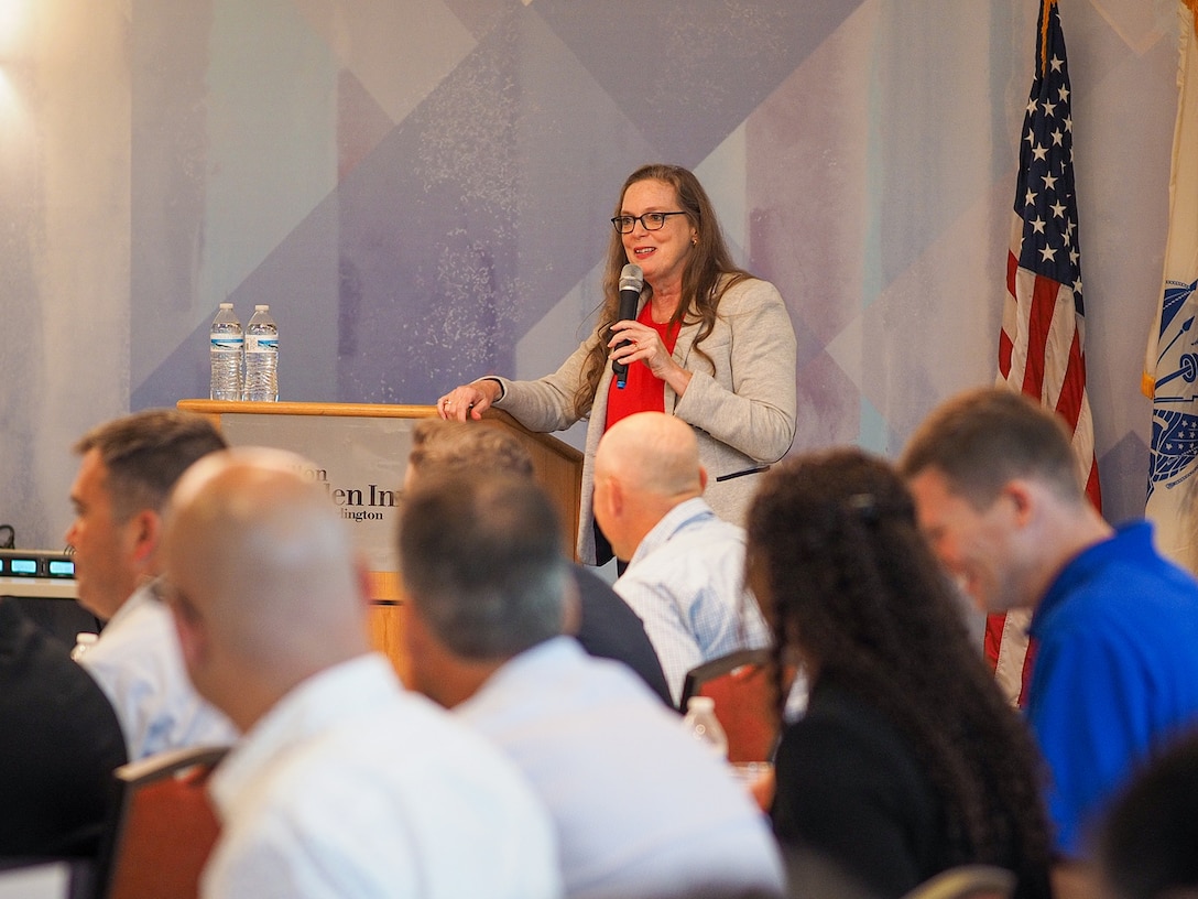 A woman with a microphone speaks to people seated at tables in a conference room.