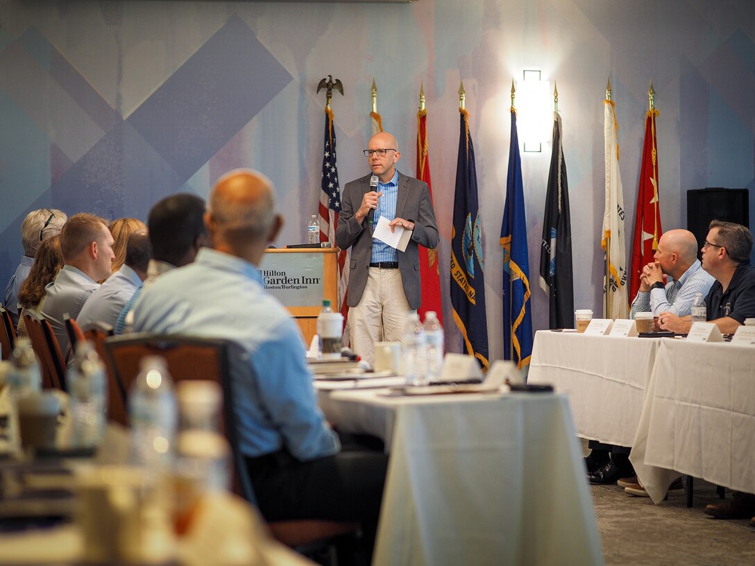 A man with a microphone talks to people seated at tables in a conference room.