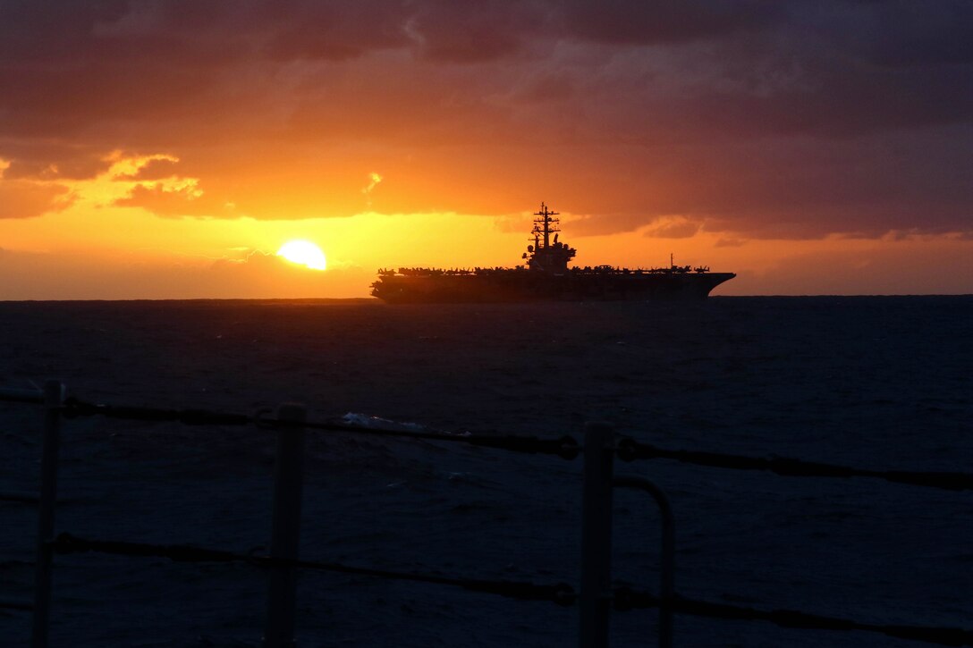 An aircraft carrier is seen from the rails of a second ship at twilight.