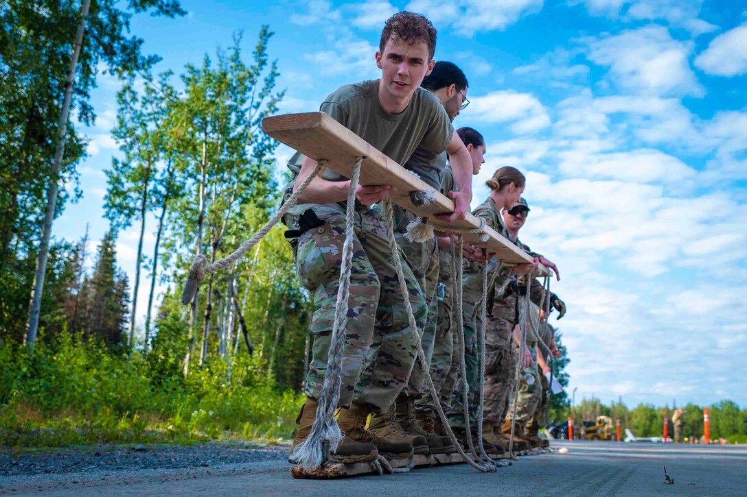 Airmen stand on a wooden plank while holding another wooden plank with ropes attached.