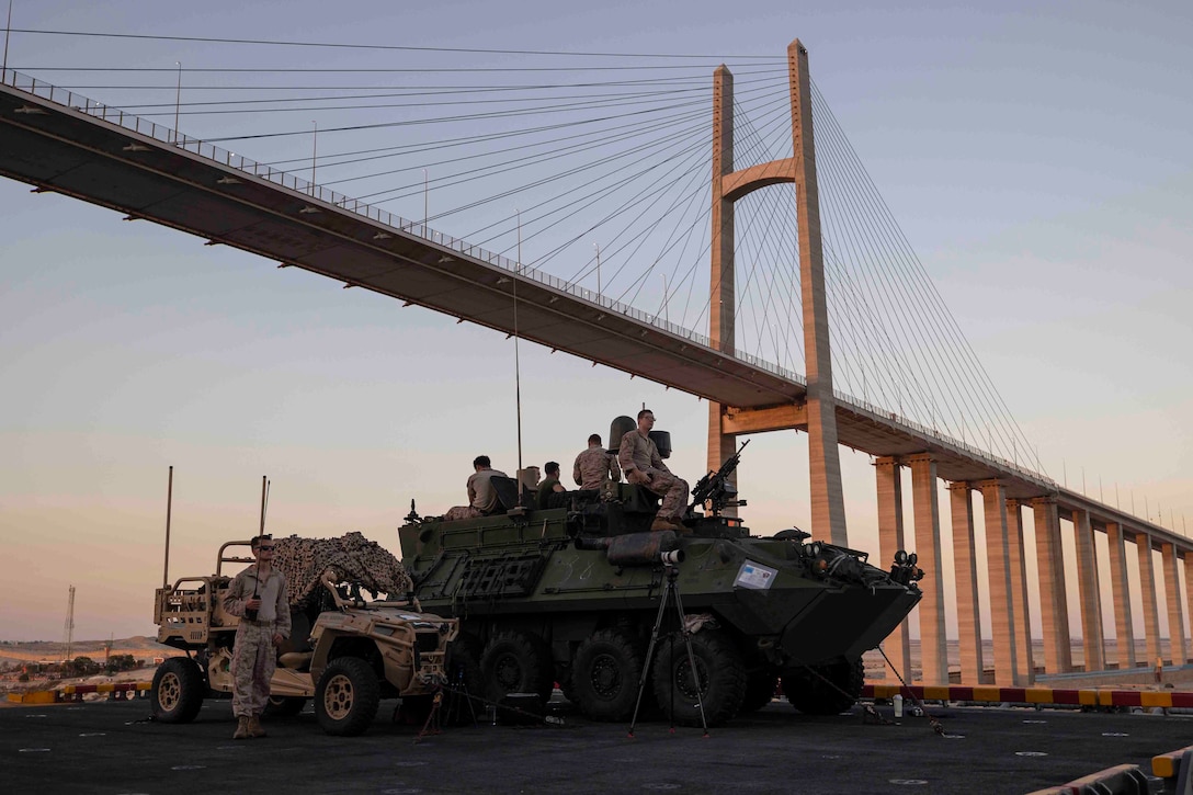 Marines and sailors sit on top of a military vehicle while another service member stands beside them next to a smaller vehicle on the deck of a ship while the ship passes under a large bridge.