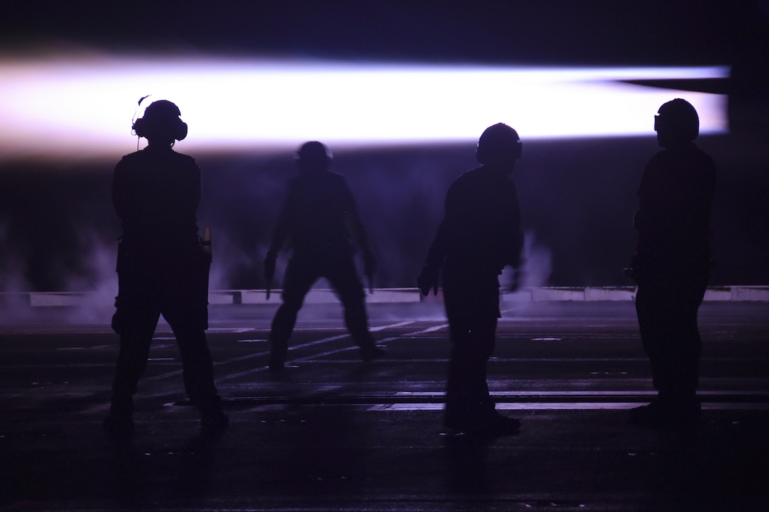 Sailors wearing headphones stand next to one another on the flight deck of a ship in the dark illuminated by a white light in the background.