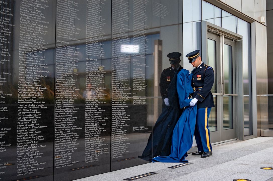 A soldier holds a drape next to a wall of engraved names.