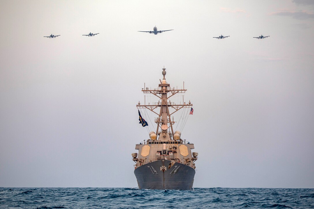 A Navy ship sails as five military aircraft fly above it.