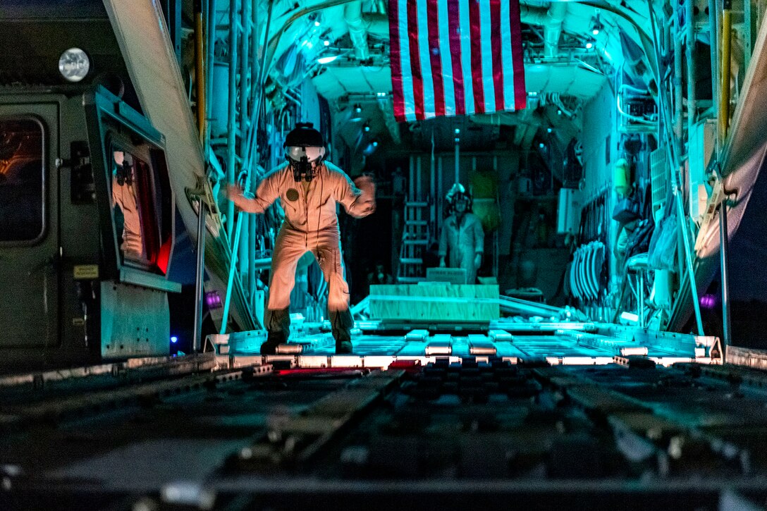 A uniformed Marine signals while standing inside a military aircraft. The read of the aircraft is illuminated in blue.