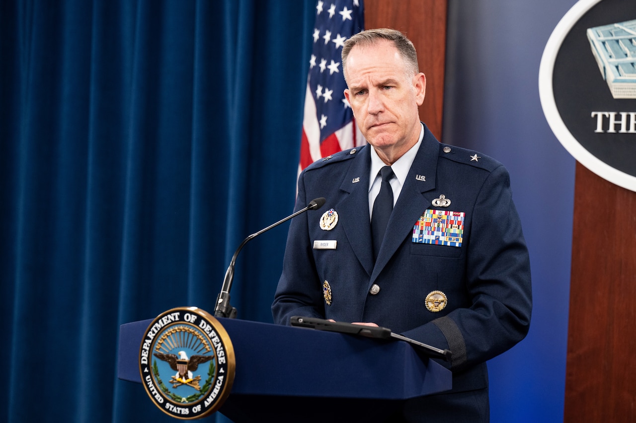 A man in uniform speaks to a group from behind a lectern with the Department of Defense seal.