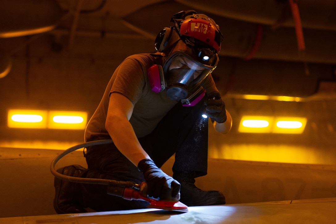 A sailor wearing a visor and an oxygen mask works on an aircraft.