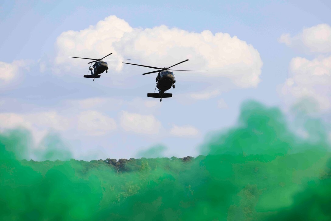 Two helicopters fly over a wooded area as green smoke rises below.