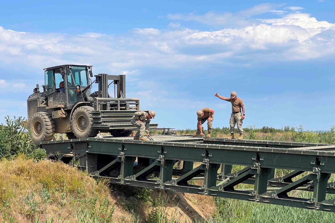 A sailor uses a forklift to carry bridge decking as three other sailors work on a bridge.