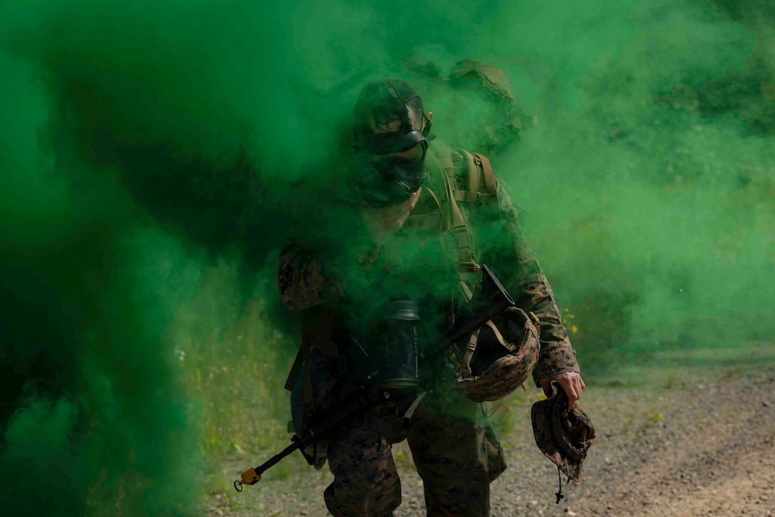 A Marine wearing a gas mask walks through a haze of green smoke.