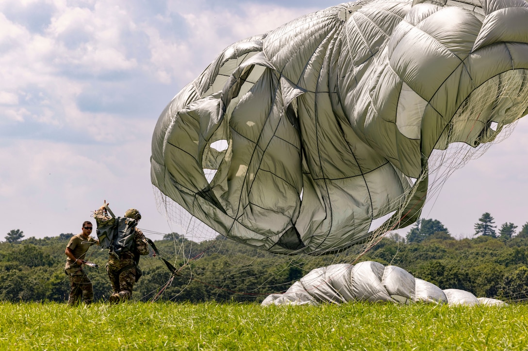 A parachute descends as a parachutist lands on the grass while speaking to a service member carrying a clipboard.