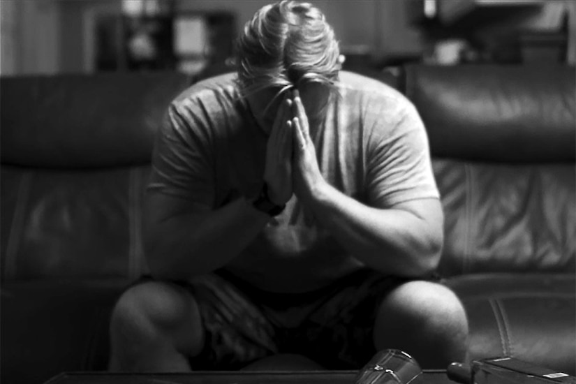 A man sits on a couch with his head pressed into his hands.