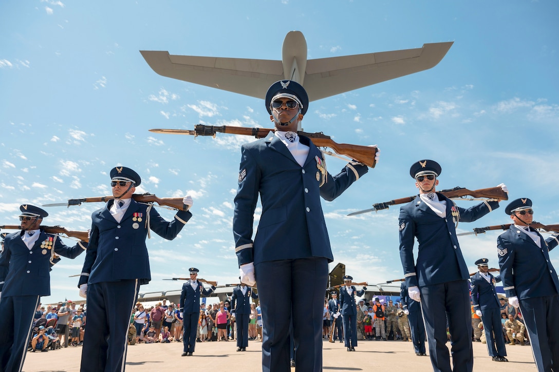 Airmen in ceremonial uniform perform with rifles in front of a crowd and a display of aircraft as an aircraft flies above.