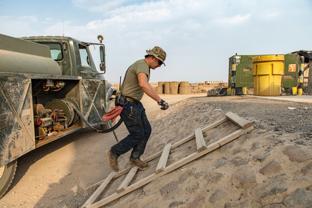 An airman carrying a fuel hose from a truck walks up a wooden ladder on a hill with containers in the background.