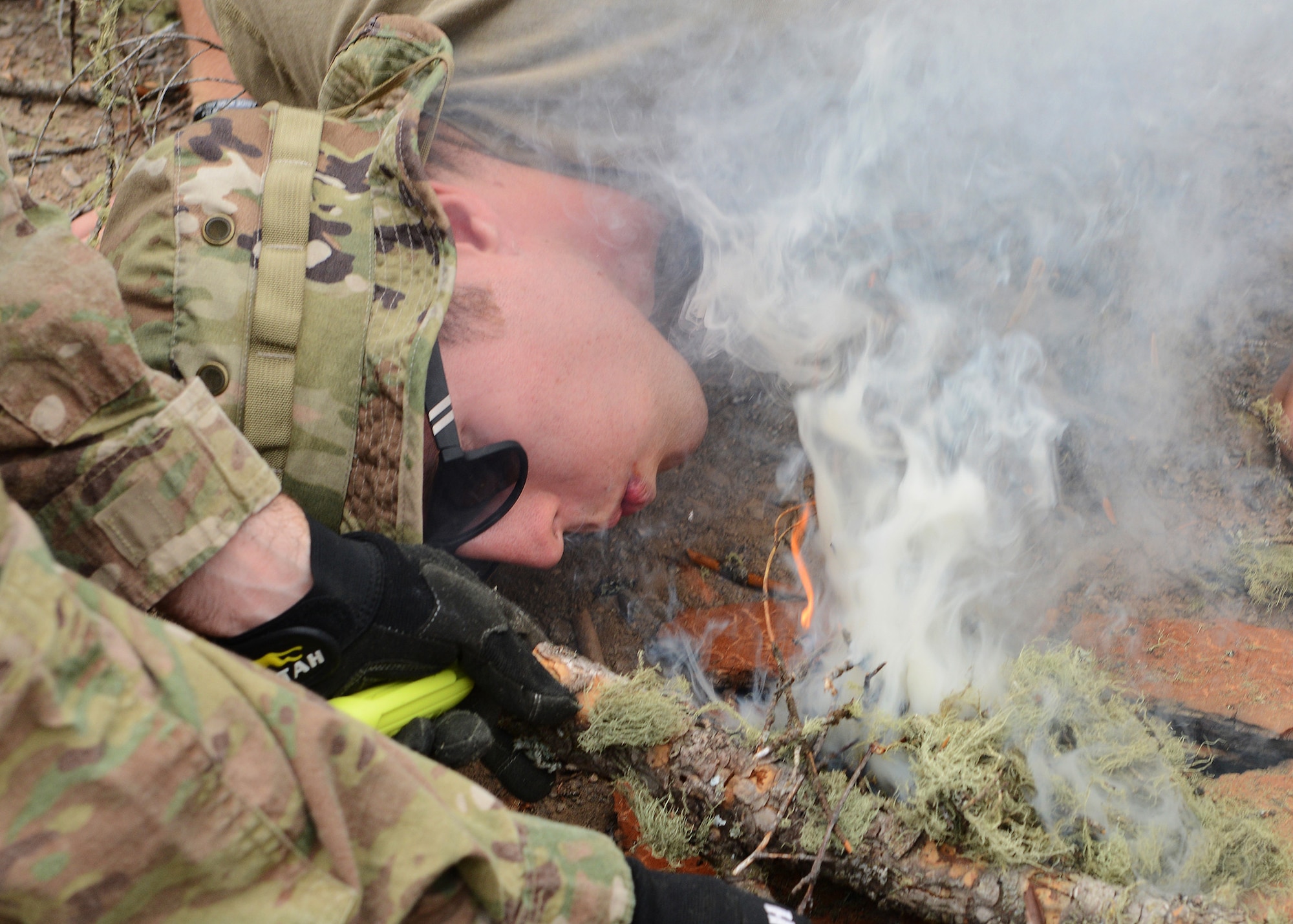 A man wearing sunglasses blows on smoky embers to create a fire.