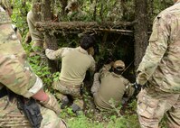 Two men kneel between a tree as they build a shelter from branches and pine boughs.