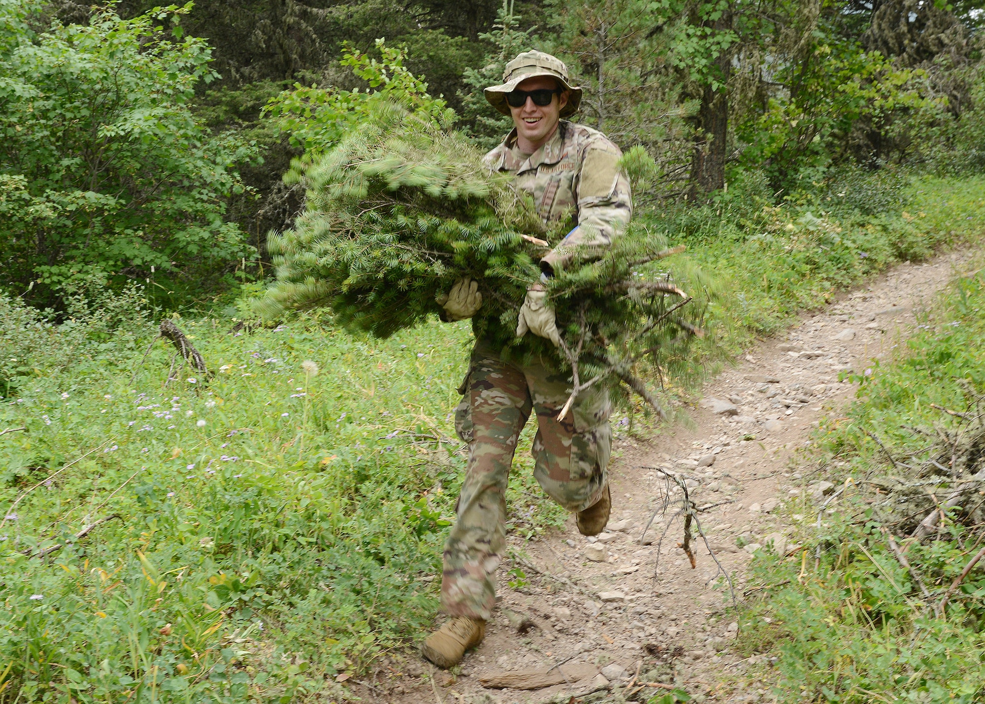 A man in military uniform runs towards the camera with arms full of pine boughs.