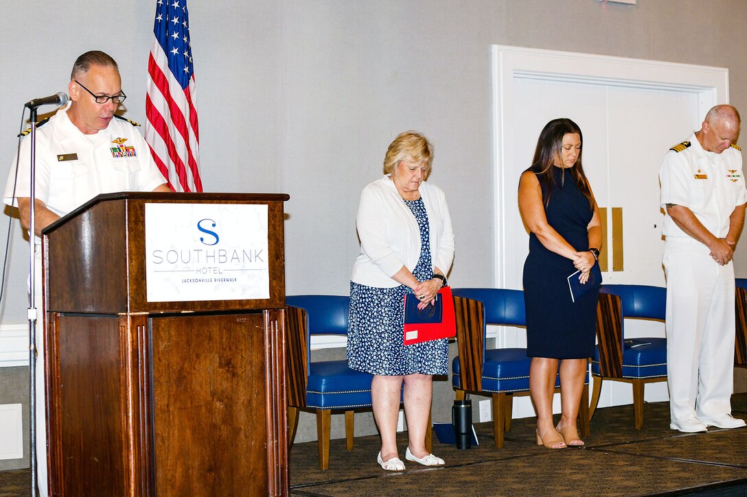 Cmdr. Donald W. Rogers Jr., presiding chaplain, delivers the invocation during Fleet Readiness Center Southeast’s (FRCSE) 2023 apprenticeship graduation ceremony at Southbank Hotel Jacksonville Riverwalk. FRCSE’s Production Support Branch Head Mary Ann Ball, MRO Production Director Veronica Amato and Commanding Officer Capt. Al Palmer bow their heads in prayer. FRCSE launched the renewed apprenticeship program in 2019, a combination work-study program that provides apprentices with a competitive wage and benefits while learning a journey-level trade. (U.S. Navy Photo by Toiete Jackson/Released)