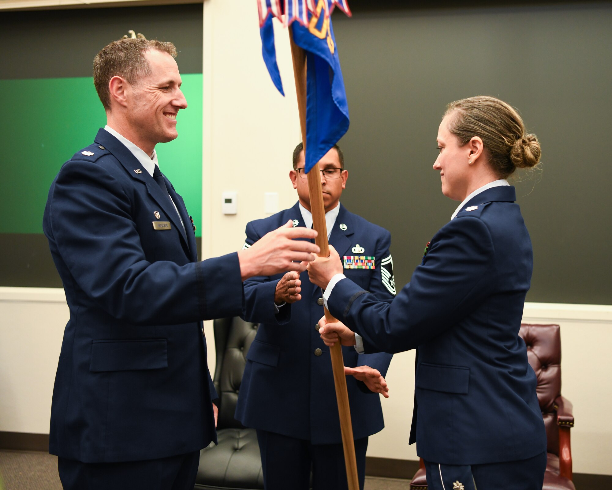 A smiling Airman in his dress blue uniform hands a blue unit flag to a female Airman during a ceremony in a classroom