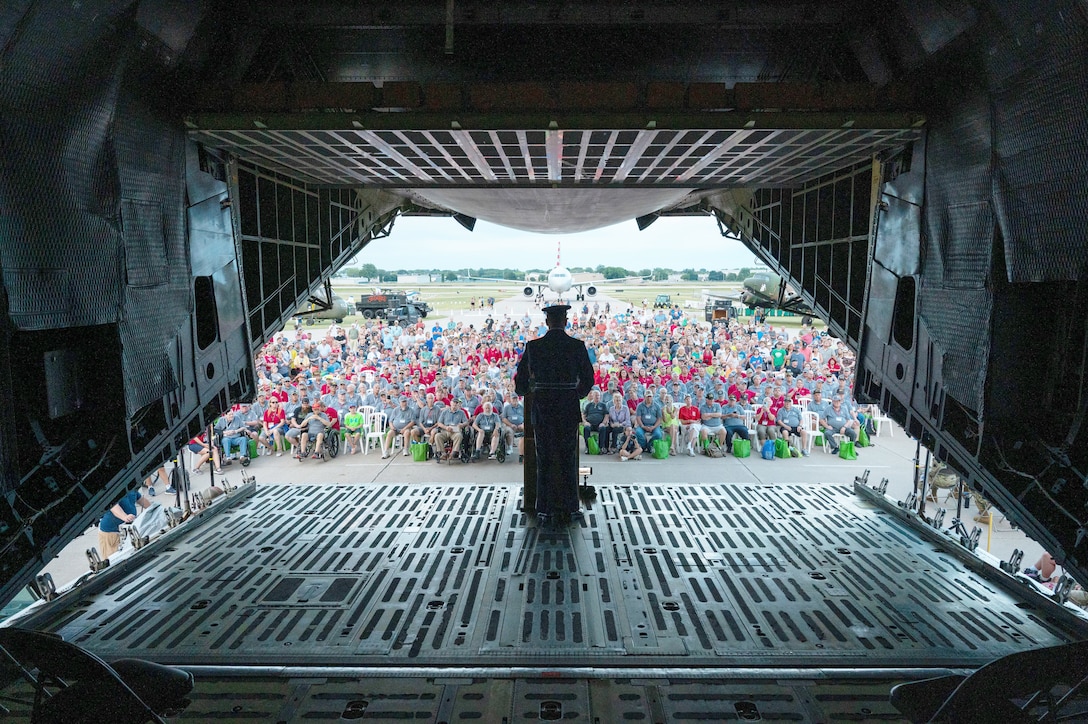 An airman stands in the loading dock of a large aircraft  and speaks to a crowd seated outside.