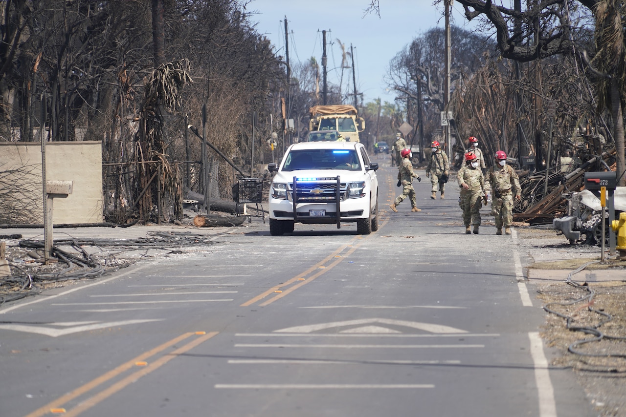 An emergency vehicle drives down a street as service members walk alongside it, assessing wildfire damage.