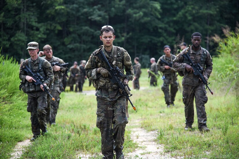 Soldiers cross a field while holding weapons.