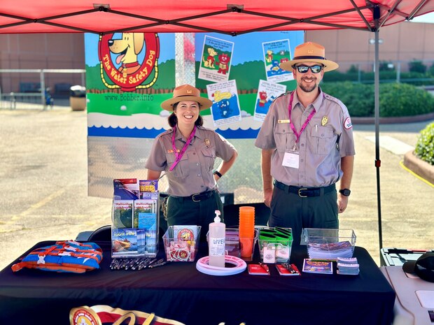 a man and a woman in park ranger uniforms are posed in front of a trailer and behind a table under a canopy