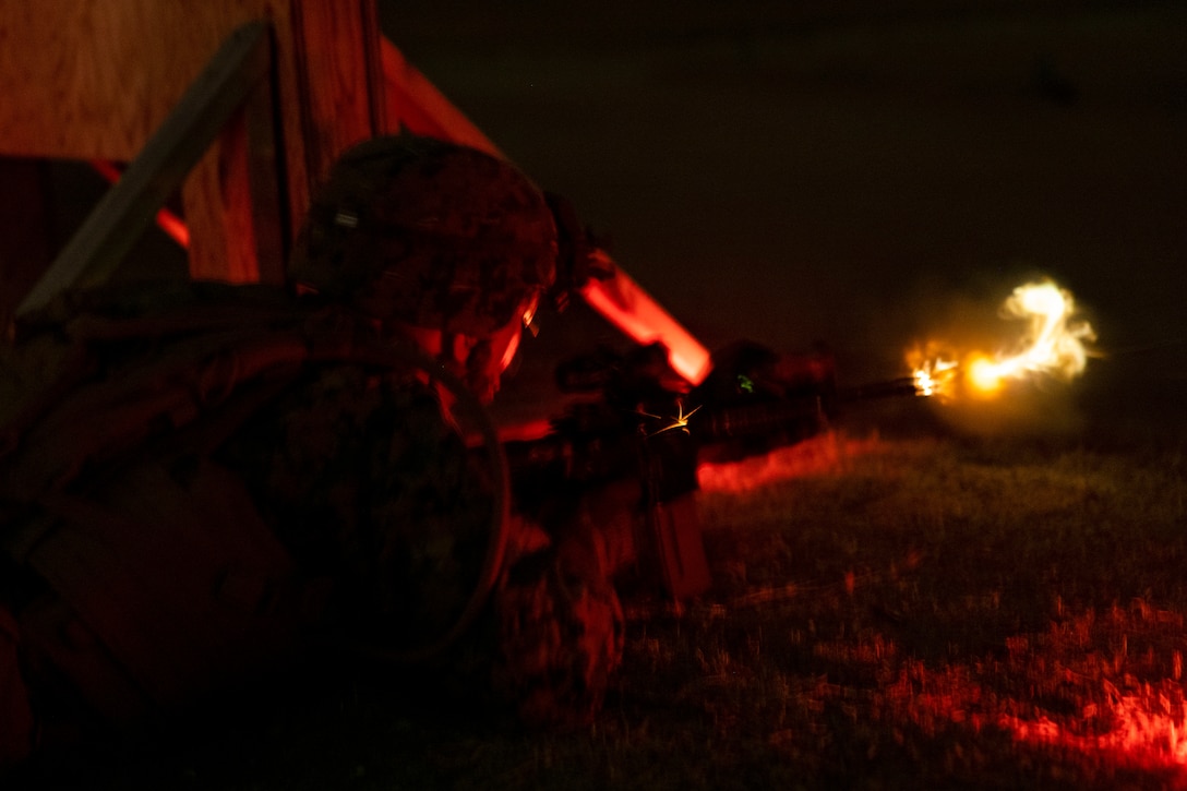 A Marine laying on the ground in the dark near a wood structure fires a weapon illuminated by orange light.