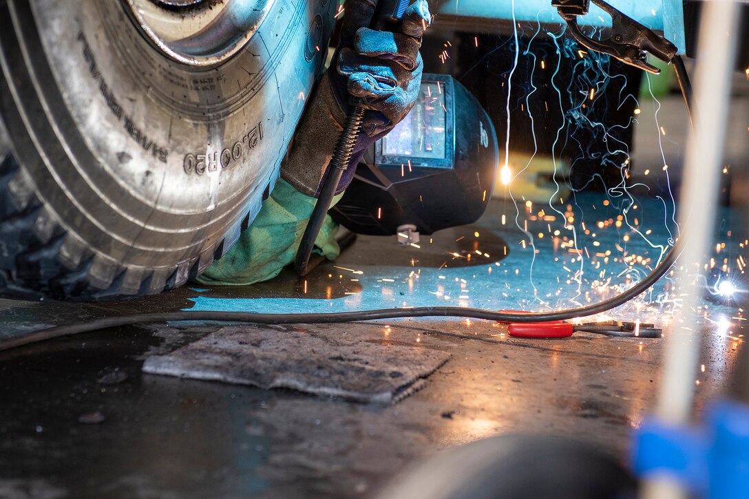 A National Guardsman lies on the floor to perform maintenance on a military vehicle as sparks emit from a tool.