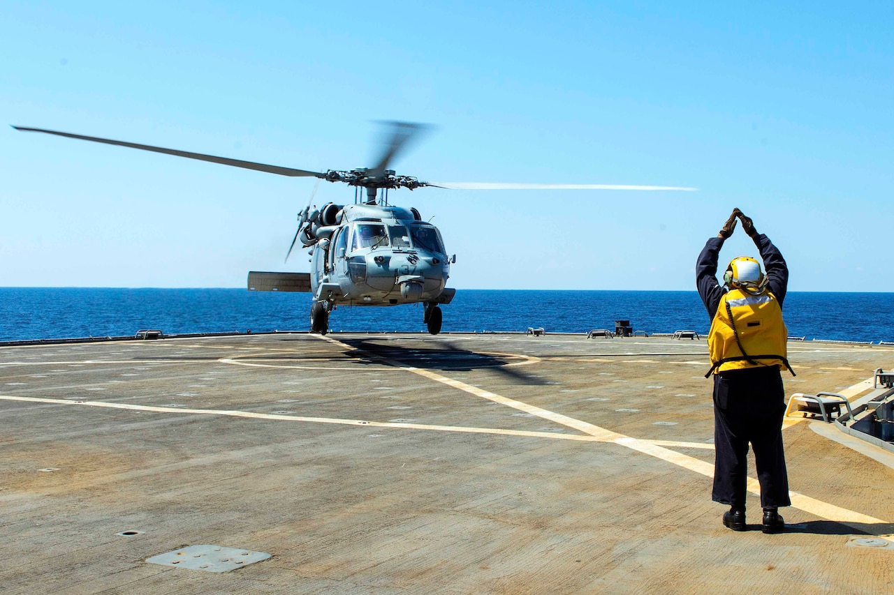 A sailor signals toward a helicopter on the deck of a ship.