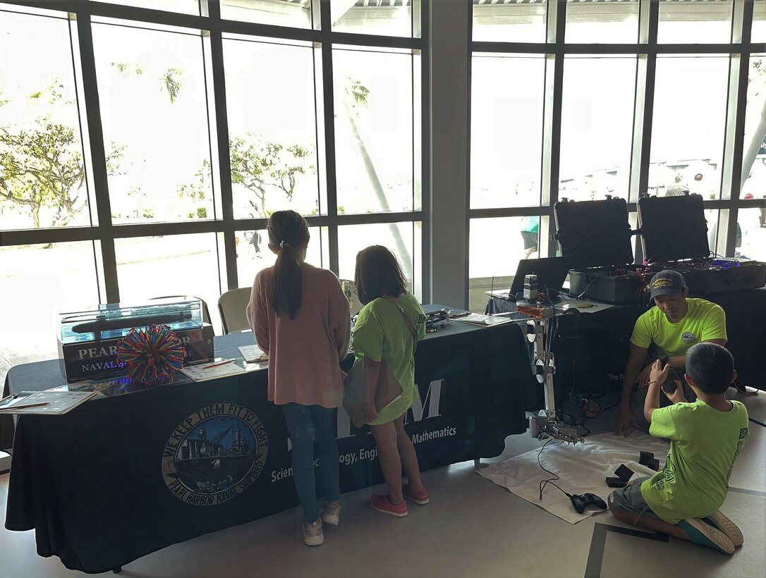 Pearl Harbor Naval Shipyard (PHSNY) Engineer Vance Hashimoto, right, from Code 100TO.32 Technology Insertion, listens to a question from a student operating a robotic arm, as additional students, left, view the demonstrations at the table, during a STEM event at the Pacific Fleet Submarine Museum.