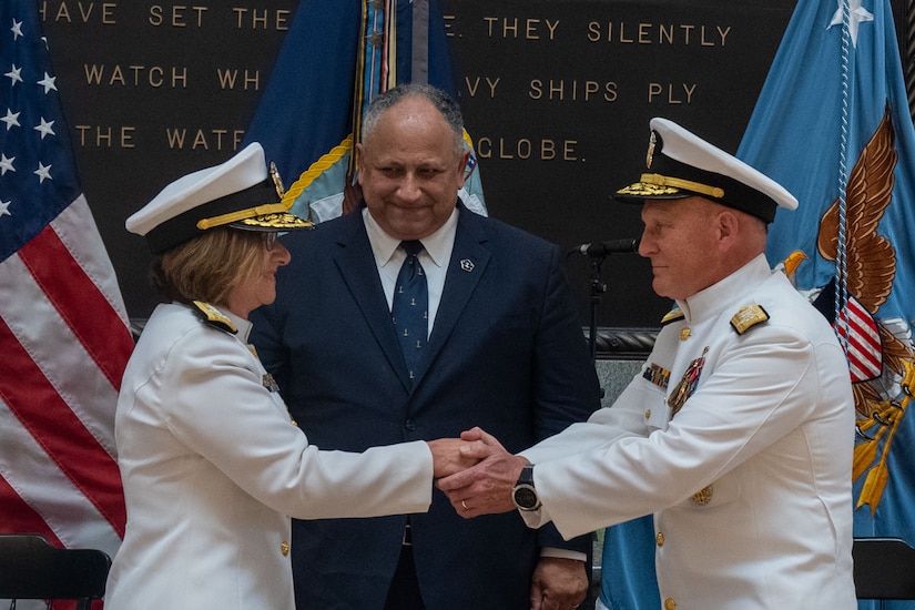 A woman and man in uniform shake hands as a man in a suit looks on.