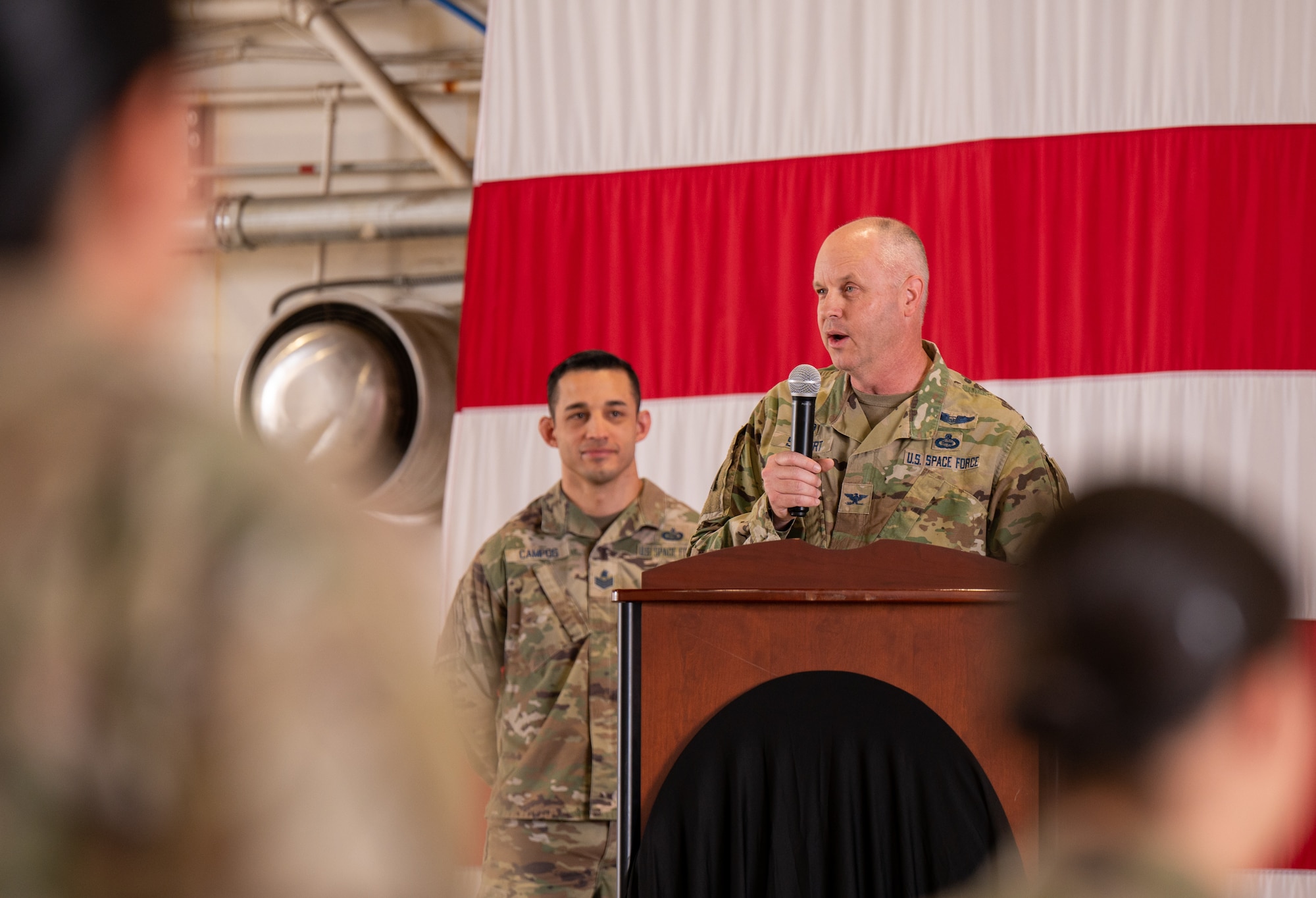 U.S. Space Force Col. Brett Swigert, Space Delta 7 - Intelligence, Surveillance and Reconnaissance commander, on stage with a large American flag behind him giving a speech to the 75th ISRS squadron