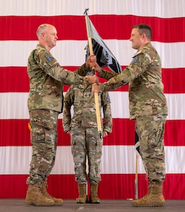 A photo of U.S. Space Force Col. Brett Swigert, Space Delta 7 - Intelligence, Surveillance and Reconnaissance commander, on a stage with a large American flag in the background, handing the ceremonial guidon to Lt. Col. Travis Anderson, 75th Intelligence, Surveillance and Reconnaissance Squadron commander