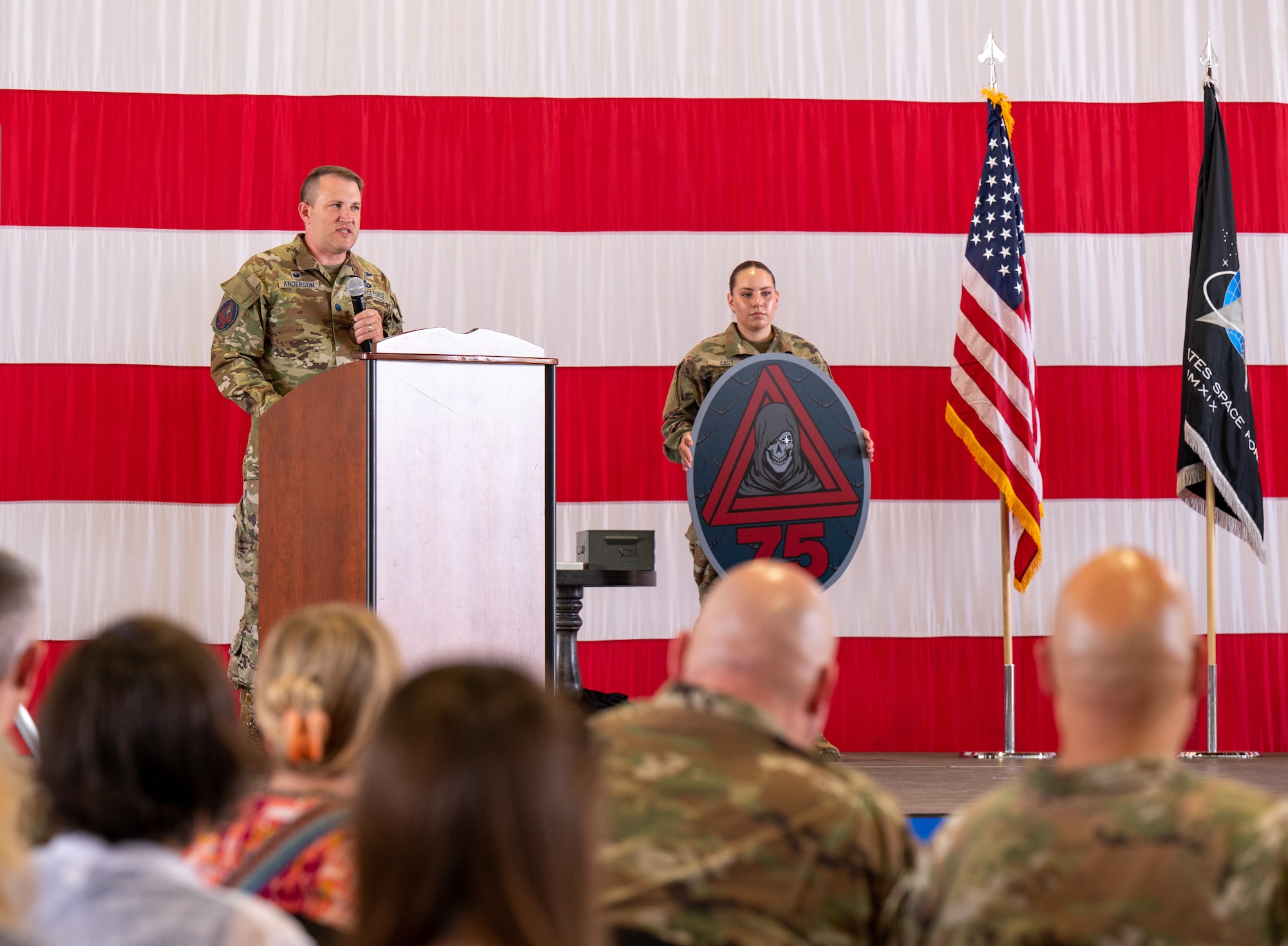 A photo of U.S. Space Force Lt. Col. Travis Anderson, 75th Intelligence, Surveillance and Reconnaissance Squadron commander, on a stage with a large American flag behind him, giving a speech.
