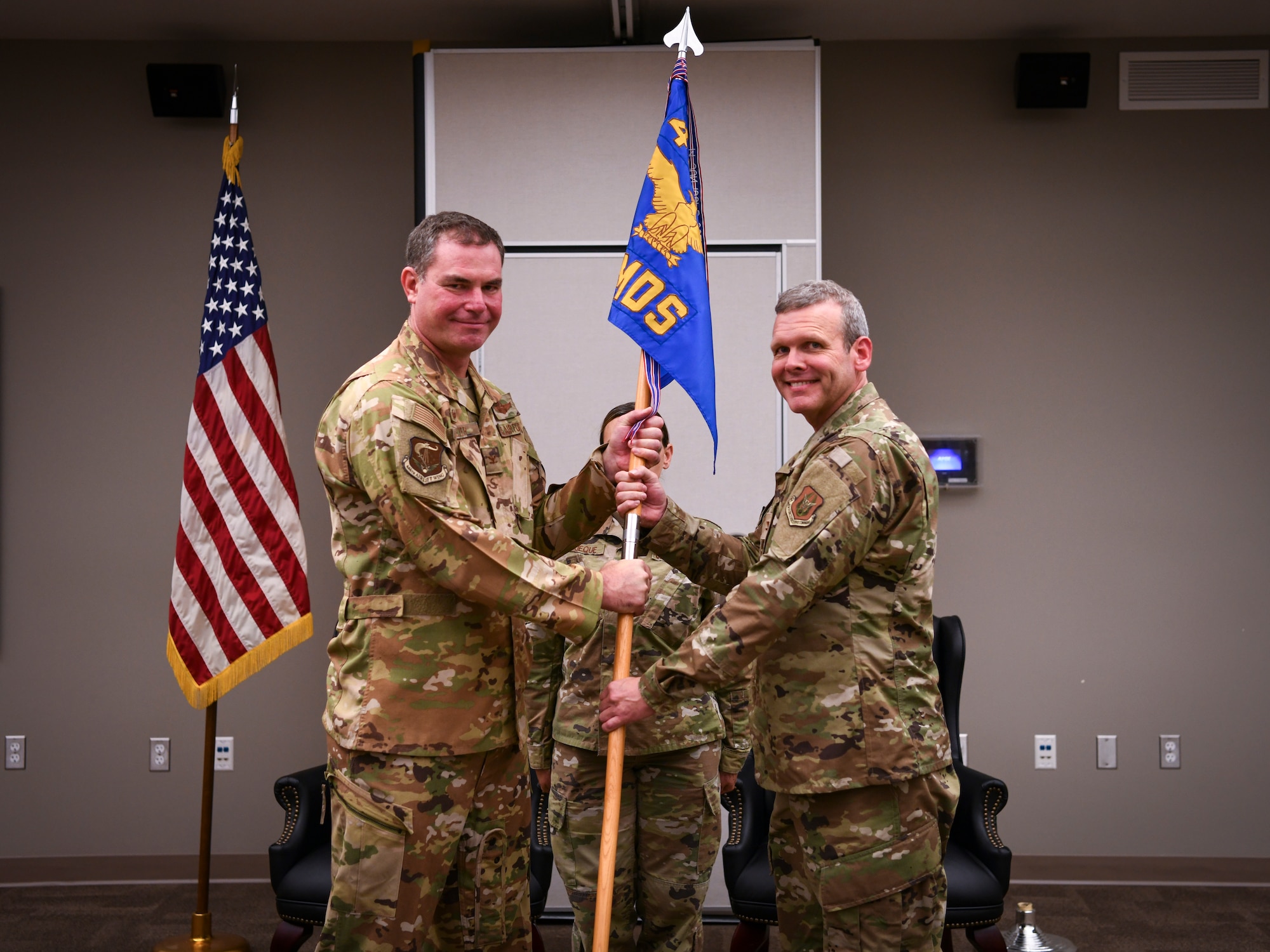 Two men in camo pattern uniforms pose with a blue guidon flag during a ceremony.