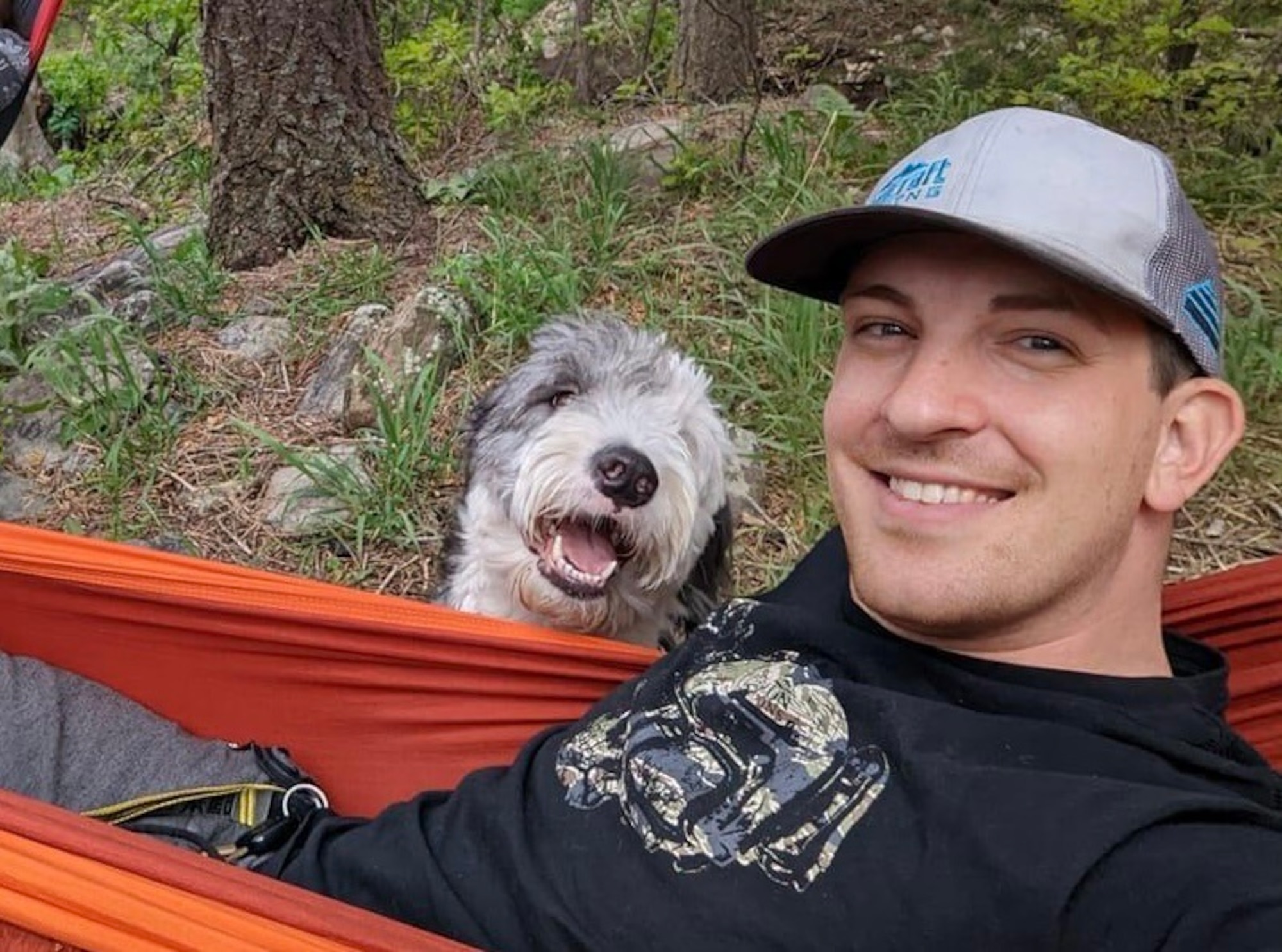Young man poses for a selfie in a hammock with a shaggy grey and white dog