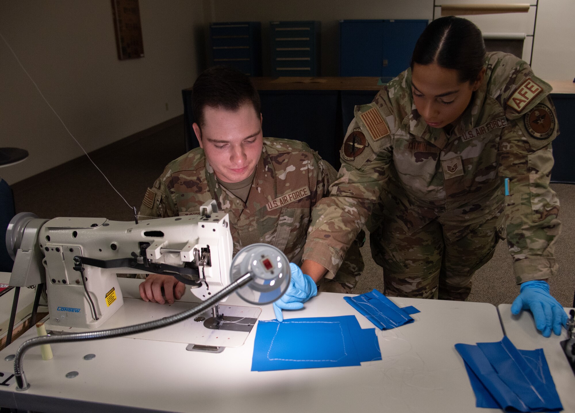 An Air Force instructor leans over an Airman at a sewing machine