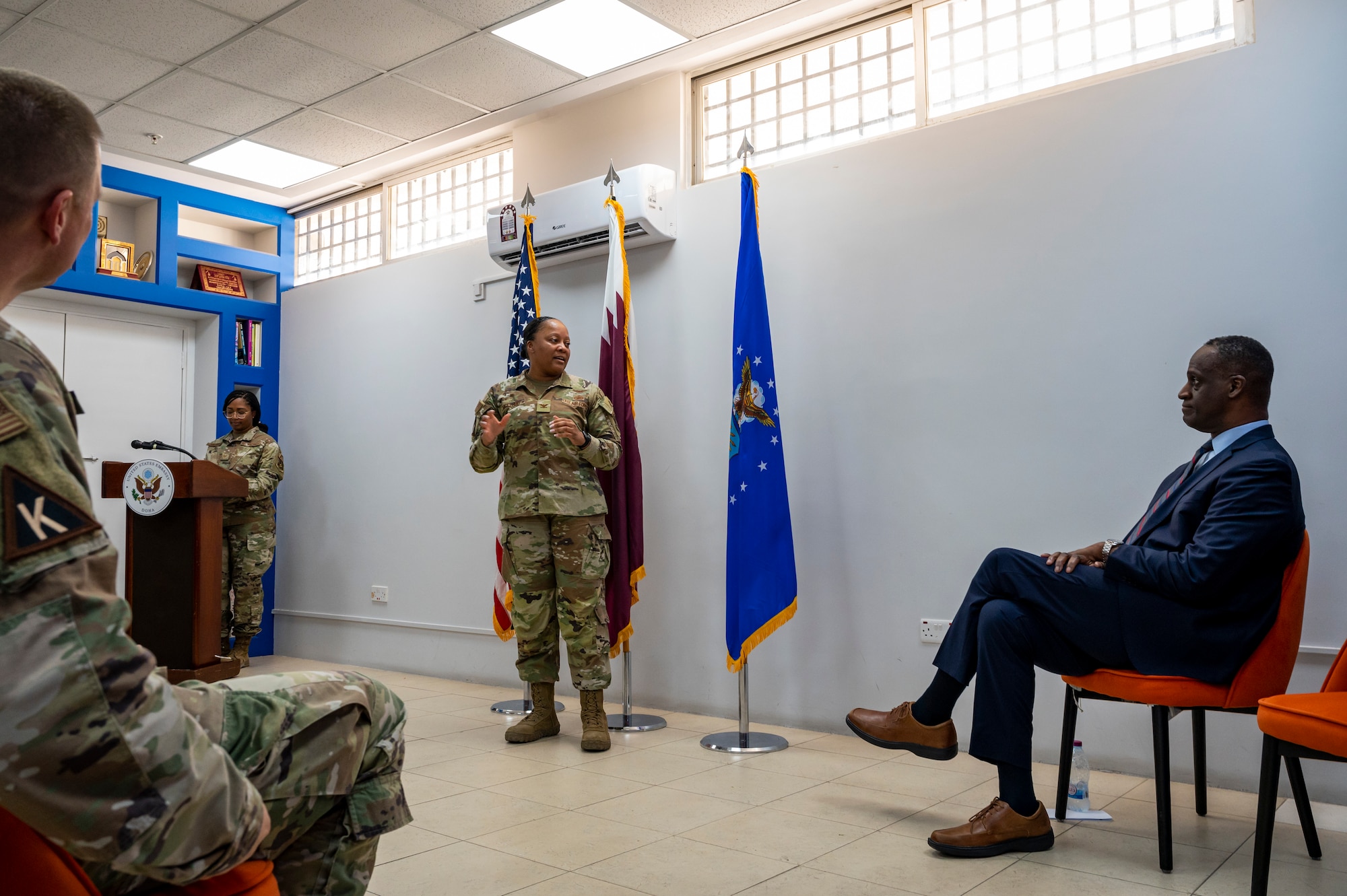 A military member in uniform talks to a crowd and the U.S. Ambassador to Qatar as they sit in the audience.