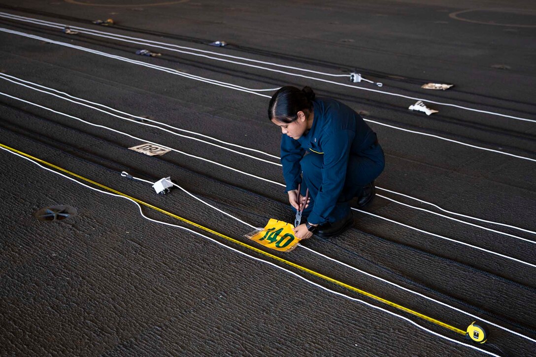 A sailor uses a tool on a piece of paper taped to the floor while surrounded by ropes lined on the floor.