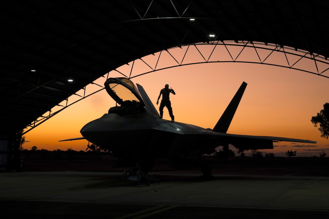 An airman stands on an aircraft in a hangar with an orangish sky in the background as shown in silhouette.