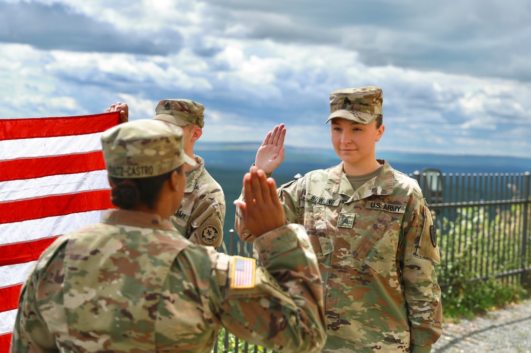 Two soldiers face each other while holding up their right hands as a third soldier holds an American flag to the left.