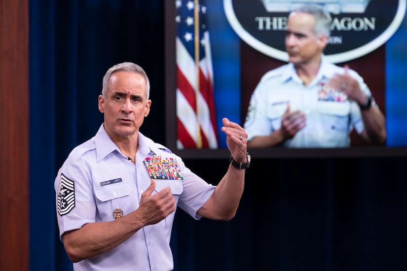 A man in uniform gestures with his hands as he speaks in front of a screen.