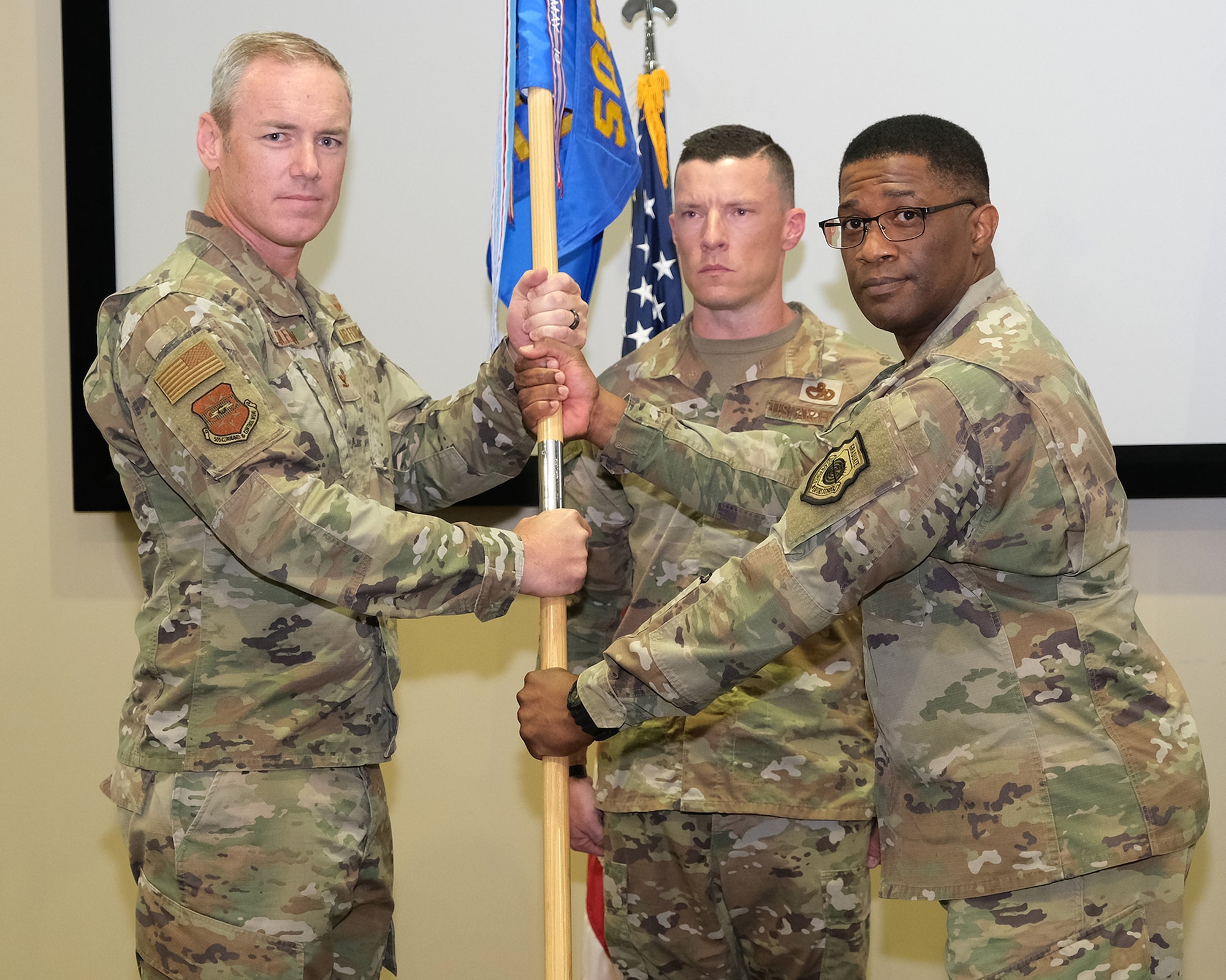 photo of three uniformed US Air Force Airmen standing on a stage, two Airmen in the forefront are both holding a unit flag while third Airman stands in background at attention