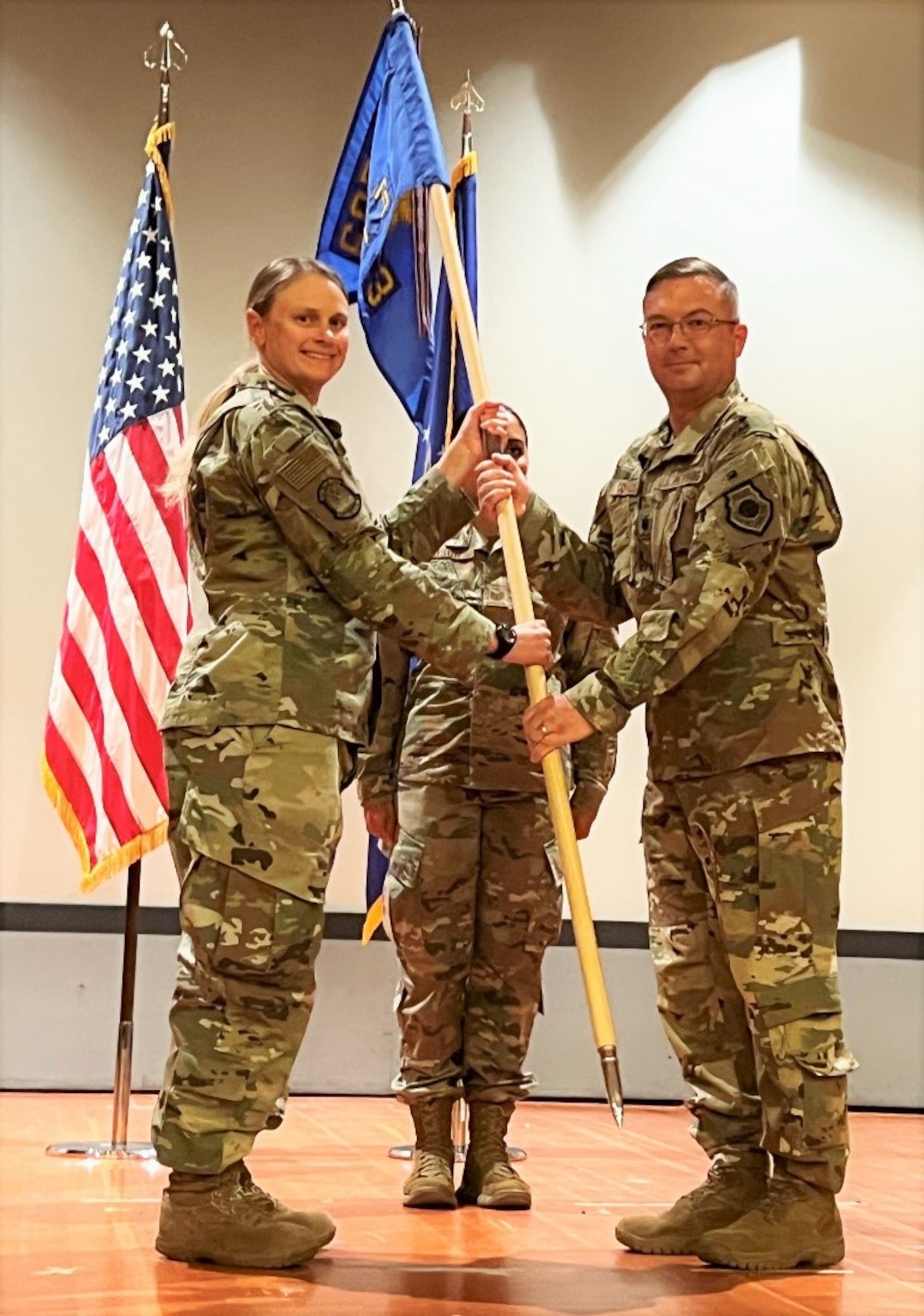 photo of three uniformed US Air Force Airmen standing on a stage, two Airmen in the forefront are both holding a unit flag while third Airman stands in background at attention