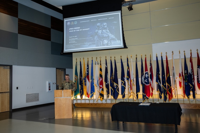 A military leader is dressed in fatigues as he addresses an audience at a podium. Presentation slides are projected over his head and he is flanked with flags to his right.
