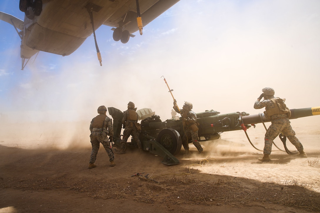 A Marine Corps helicopter hovers over Marines and a howitzer in a dusty area.